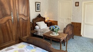 A bedroom with a large wooden wardrobe, a bed with a patterned bedspread, a bedside table with a vase of flowers, and a nightstand with a lamp. - Le Petit Gîte du Theil Au Servant (LA CHAPELLE-VIVIERS)