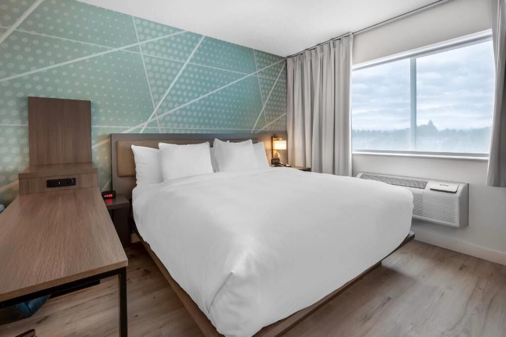 A hotel room with a large bed, a bedside table, a nightstand, a wall-mounted heater, and a window with curtains.