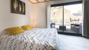 A bedroom with a bed, a large window, and a view of a pool and buildings. - Le Gite du Marronnier (MONTAMISE)