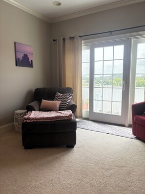 Ein Wohnzimmer mit einem dunklen Sofa, einem kleinen runden Beistelltisch, einer großen Schiebeglastür, die auf einen Balkon führt, und Blick ins Grüne.