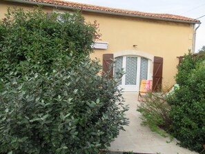 Un bâtiment avec un toit recouvert de tuiles, une porte dotée d’une fenêtre et un petit jardin agrémenté d’une chaise et de plantes.