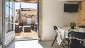 Ein offenes Durchgang führt zu einer Außenveranda mit Tisch und Stühlen, einem an der Wand befestigten Flachbildfernseher und einer weißen Decke mit Holzbalken.