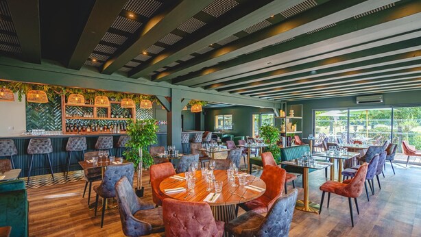 Un restaurante con mesas redondas, sillas acolchadas y una zona de bar con botellas.