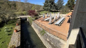 A riverside terrace with tables and chairs, a stone wall, and a bridge.