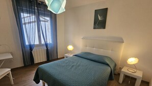 Ein Schlafzimmer mit einem Bett, zwei Nachttischen mit Lampen, einem Fenster mit Vorhängen, einem Heizkörper und einem an der Wand hängenden Bild.