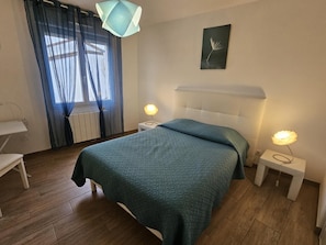 A bedroom with a bed, two bedside tables with lamps, a window with curtains, a radiator, and a wall-mounted picture. - La Blanche Maille - CISSE (CISSE)