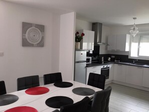 A modern kitchen with white cabinets, a stainless steel range hood, and a dining area with a white table and black chairs. - La Blanche Maille - CISSE (CISSE)