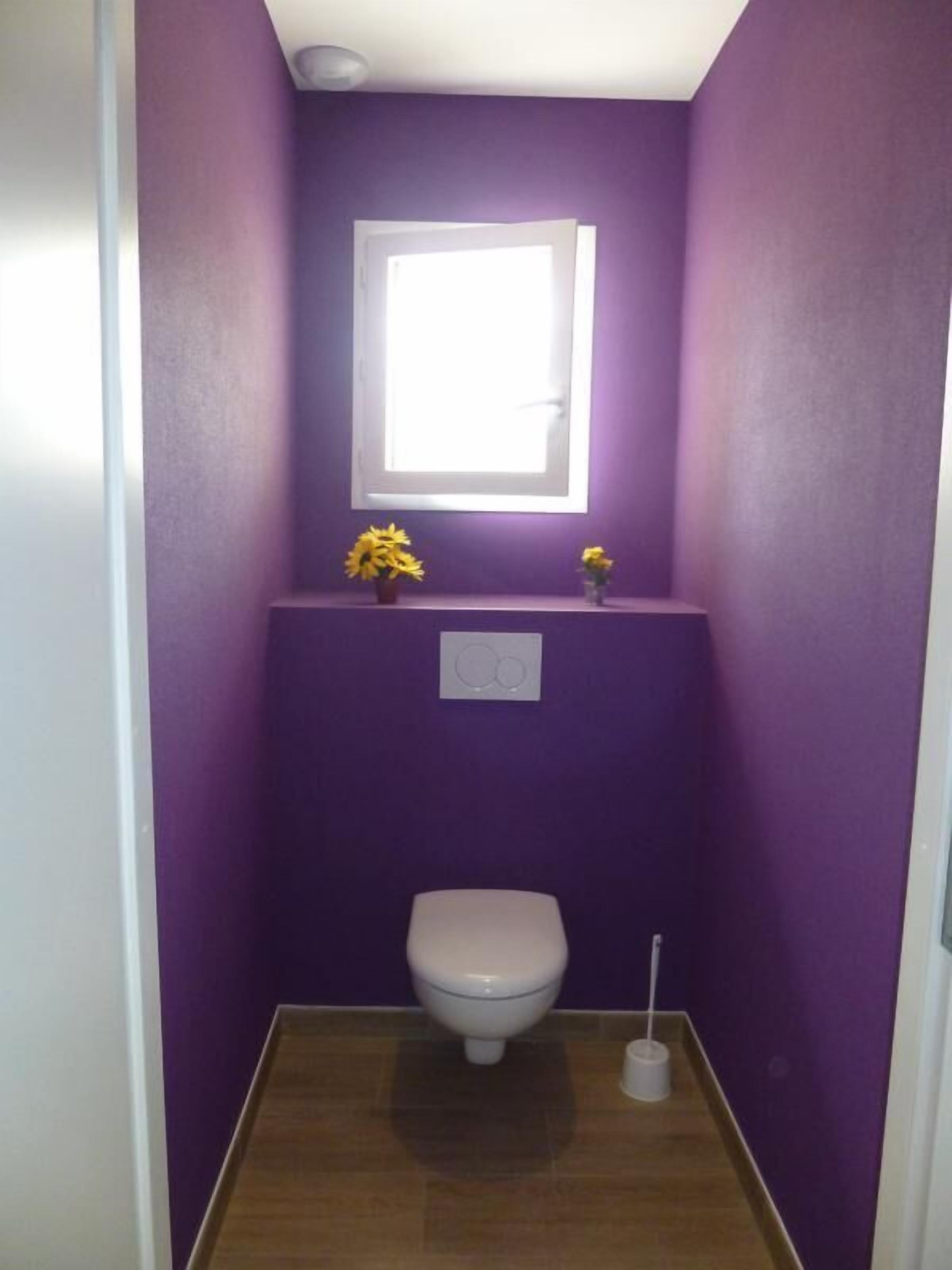 Et lille badeværelse med lilla vægge, et hvidt toilet, et vindue og en lille hylde med blomster.