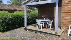 Un espace extérieur aménagé, abrité, avec une table et des chaises, une terrasse en bois et, au fond, un bâtiment dont le toit est recouvert de tuiles.