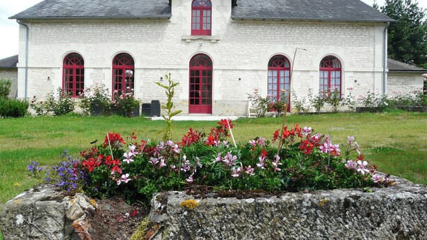 Un bâtiment blanc avec des portes et des fenêtres rouges, entouré d’un jardin où poussent différentes sortes de fleurs.