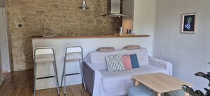 Quarto com parede de pedra, sofá branco com almofadas estampadas, mesa de madeira e cadeira de metal.