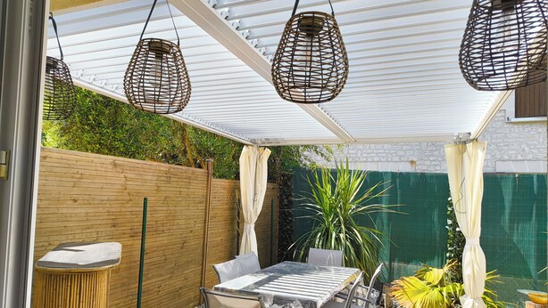 Un'area patio coperta con tavolo e sedie, lampade in vimini appese e una recinzione verde.