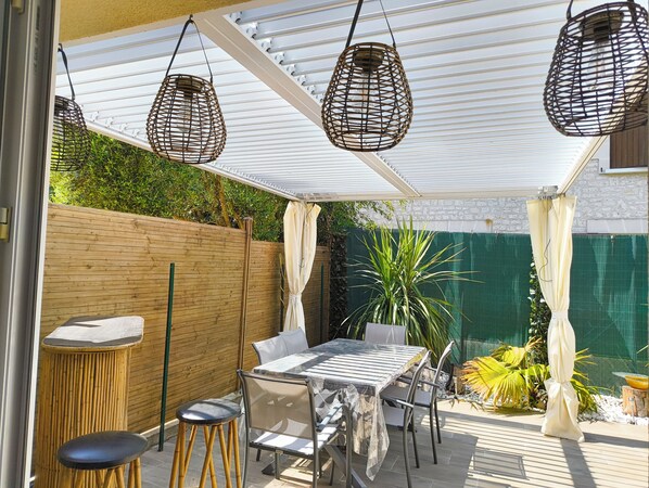 A covered patio area with a table and chairs, hanging wicker lamps, and a green fence. - La Royannaise" house by the sea (Royan)