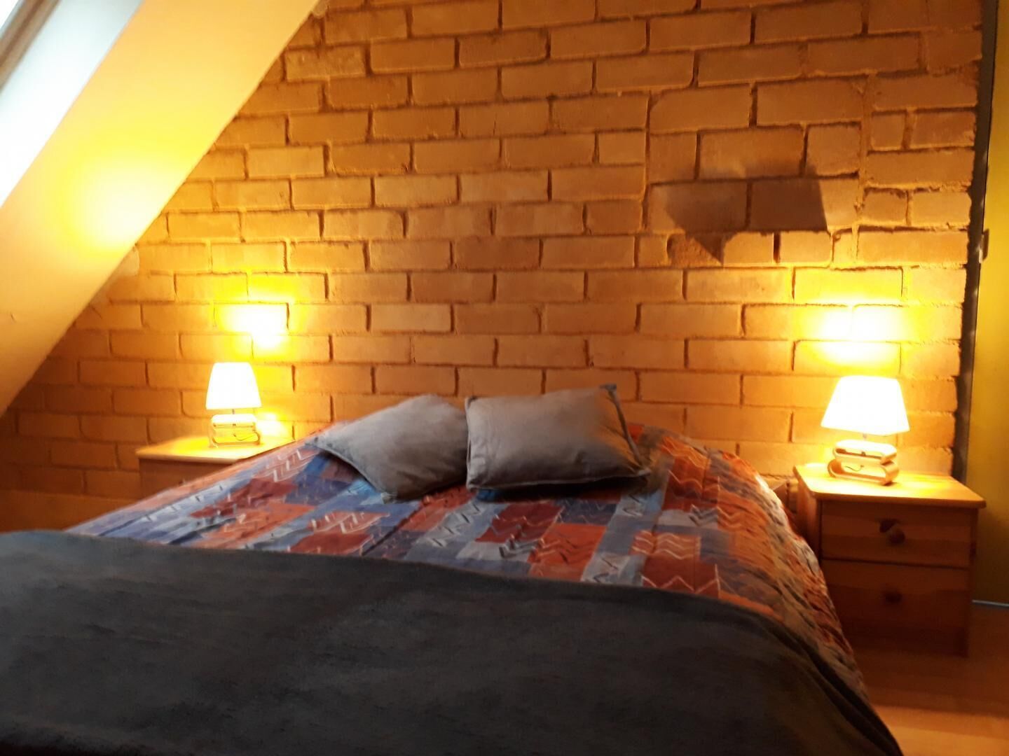A bedroom with a sloped ceiling, exposed brick wall, two bedside lamps, a bed with a patterned blanket, and two pillows.