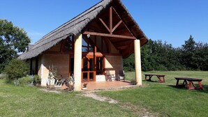 Una cabaña rústica de madera con techo de paja, un porche cubierto con asientos y una mesa de picnic en un área verde.