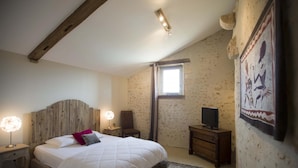 Un dormitorio con cabecera de madera, cama con ropa de cama blanca, mesita de noche con lámpara, televisor sobre un mueble de madera y ventana con vistas.