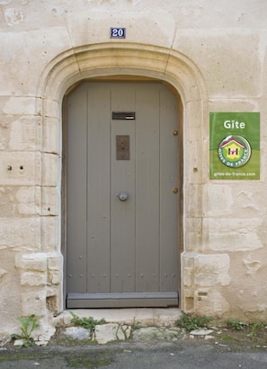 A stone building with a grey door, a number '20' sign above the door, and a green sign with 'Gîte' and a website address. - Le Clos Mélior - LUSIGNAN (LUSIGNAN)