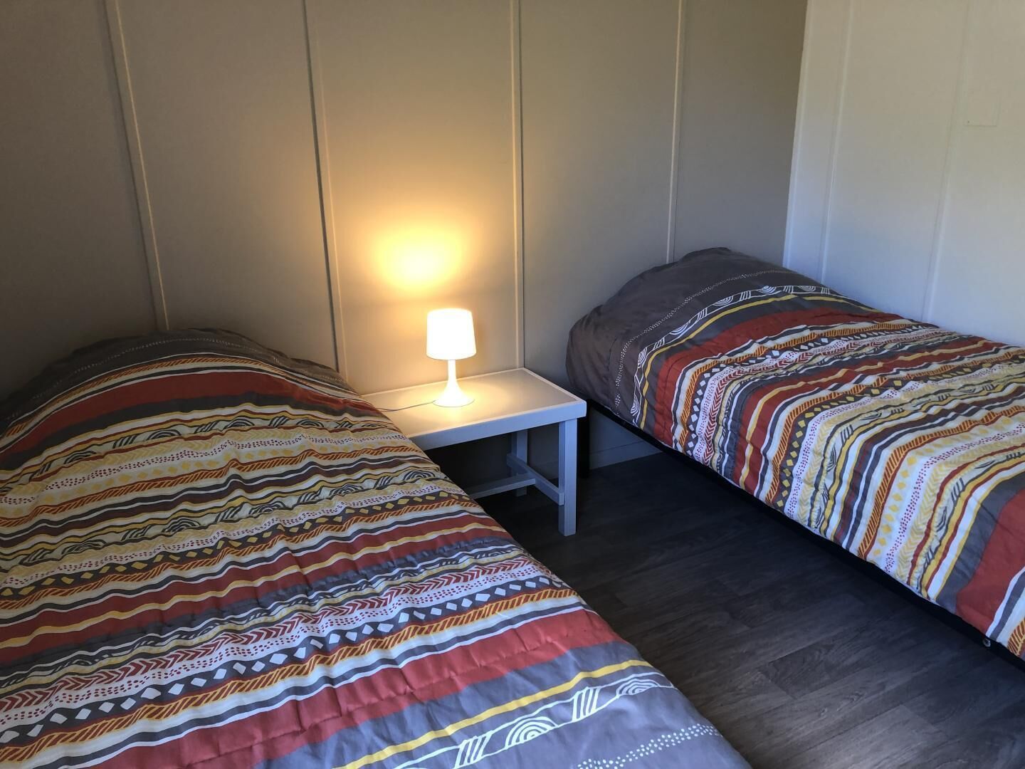 A room with two beds, a nightstand with a lamp, and a wooden floor.