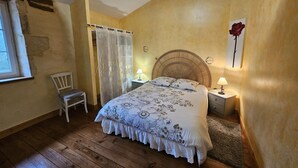 A bedroom with a wooden floor, a bed with a floral bedspread, a chair, a window with a curtain, and a wall-mounted rose decoration. - The Gallinaceans - LUSIGNAN (LUSIGNAN)