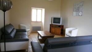 A living room with a sofa, a coffee table, a TV on a wooden stand, a radiator, and a window. - Les Cèdres - BLANZAY - Gîte (BLANZAY)