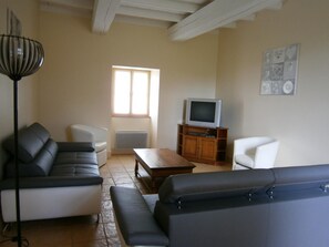 A living room with a sofa, a coffee table, a TV on a wooden stand, a radiator, and a window. - Les Cèdres - BLANZAY - Gîte (BLANZAY)
