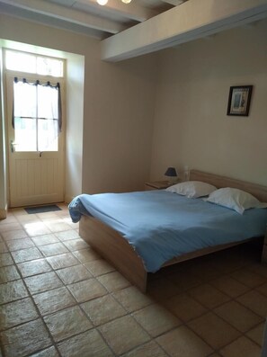 Quarto com cama, mesa de cabeceira, luminária, janela com cortinas e porta.
