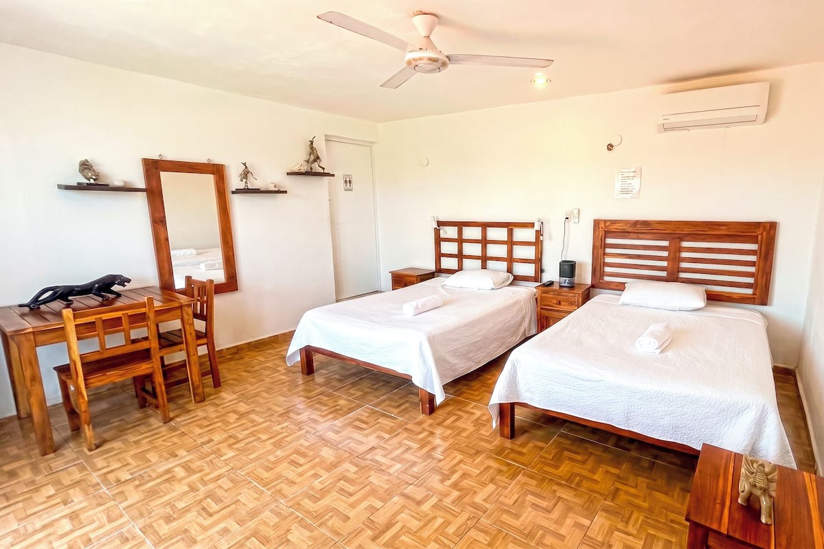 Ein Hotelzimmer mit zwei Betten, einem Holztisch, einem Stuhl, einem Deckenventilator und einer Klimaanlage.