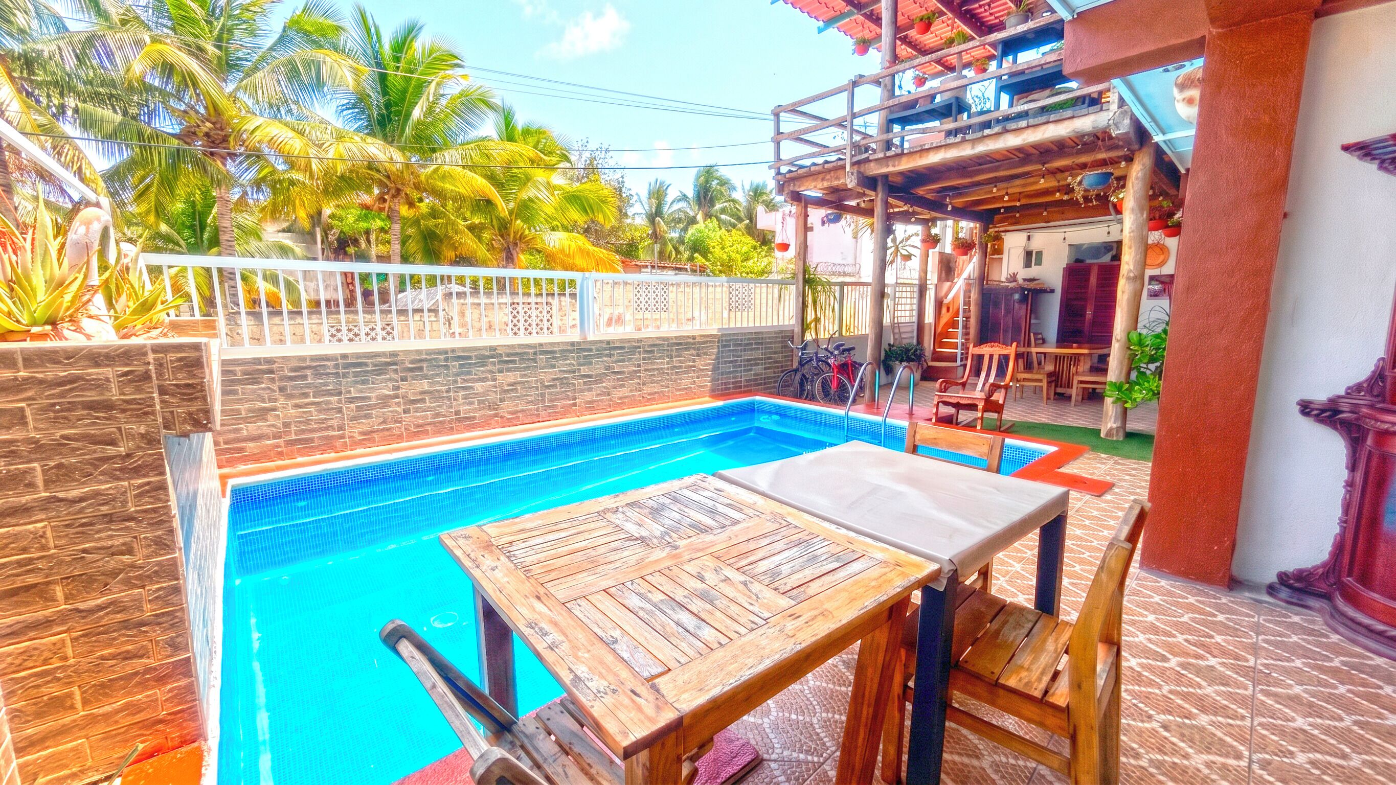 A pool area with a wooden table and chairs, surrounded by a stone wall and palm trees.