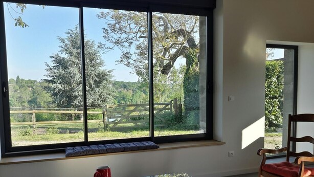 Una habitación con un ventanal grande, una mesa de vidrio con una taza roja, una mecedora de madera y un banco con cojines.