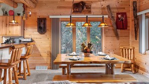 A wooden cabin interior with a dining table, chairs, pendant lights, a kitchen area, and a large window. - Rustic Ridge Cabin 104 - Private Woodland Retreat near Devil's Lake State Park (Merrimac)
