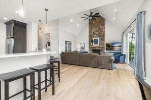 A modern kitchen with a stone accent wall, a dining area with bar stools, a living room with a sofa and fireplace, and large windows with curtains. - New!!  Lakefront, Dock, Kayaks, Firepit & Game Room (Macon)