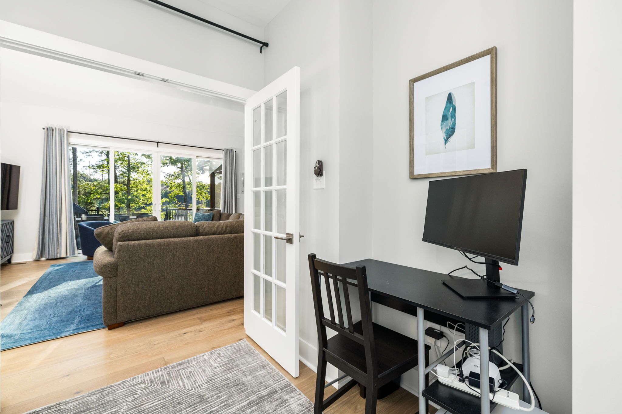 A room with a sofa, a desk with a monitor, a framed picture on the wall, and a sliding glass door leading to an outdoor area.