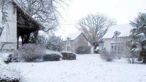 Pátio coberto de neve com edifícios de pedra, uma pérgola de madeira e diversos arbustos.