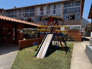 Un parque infantil con un tobogán y columpios, un edificio de ladrillo con tejado de baldosas, y un edificio residencial de varios pisos al fondo.