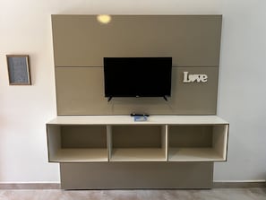 Un televisor montado en la pared con un estante debajo, un certificado enmarcado a la izquierda y la palabra 'Love' a la derecha.