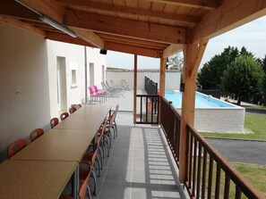 Een overdekt terras met een lange tafel en stoelen, een zwembad op de achtergrond en uitzicht op een woonwijk.