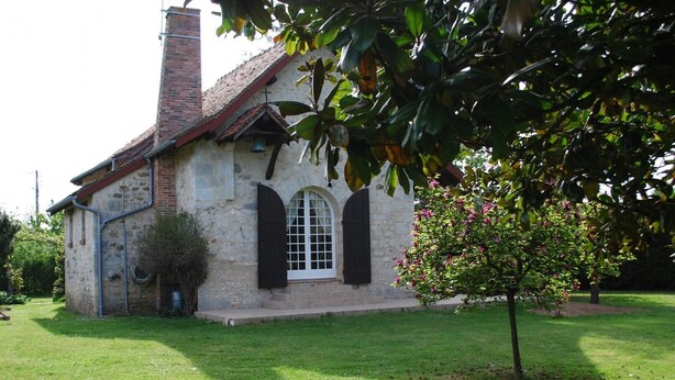 Ein Gebäude aus Stein mit Ziegeldach, einer Glocke und einem kleinen Garten mit einem Baum.