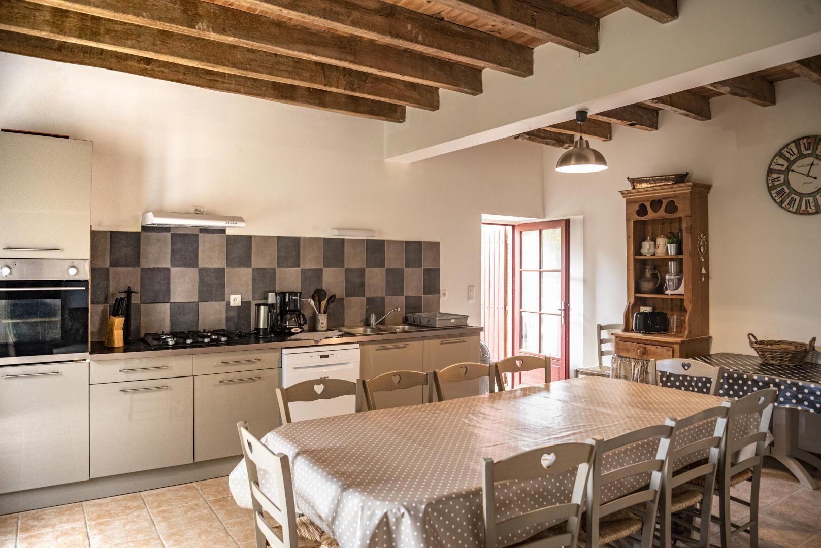 Une cuisine équipée d’une cuisinière, d’un four et d’un plan de travail à carreaux. On y trouve une table à manger avec des chaises et une horloge au mur.