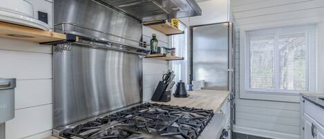 A modern kitchen with a stainless steel range hood, a gas stove, wooden shelves with kitchen items, and a window with blinds.