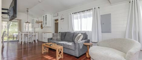A spacious living area with a grey sofa, a white armchair, a wooden coffee table, and a TV mounted on the wall.