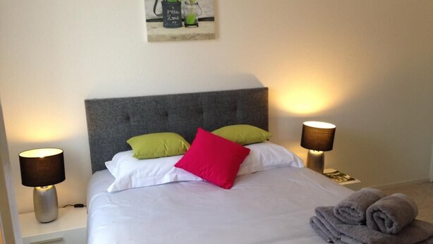 Een keurig opgemaakt bed met kussens, een bedlampje en een opgerolde handdoek op de vloer.