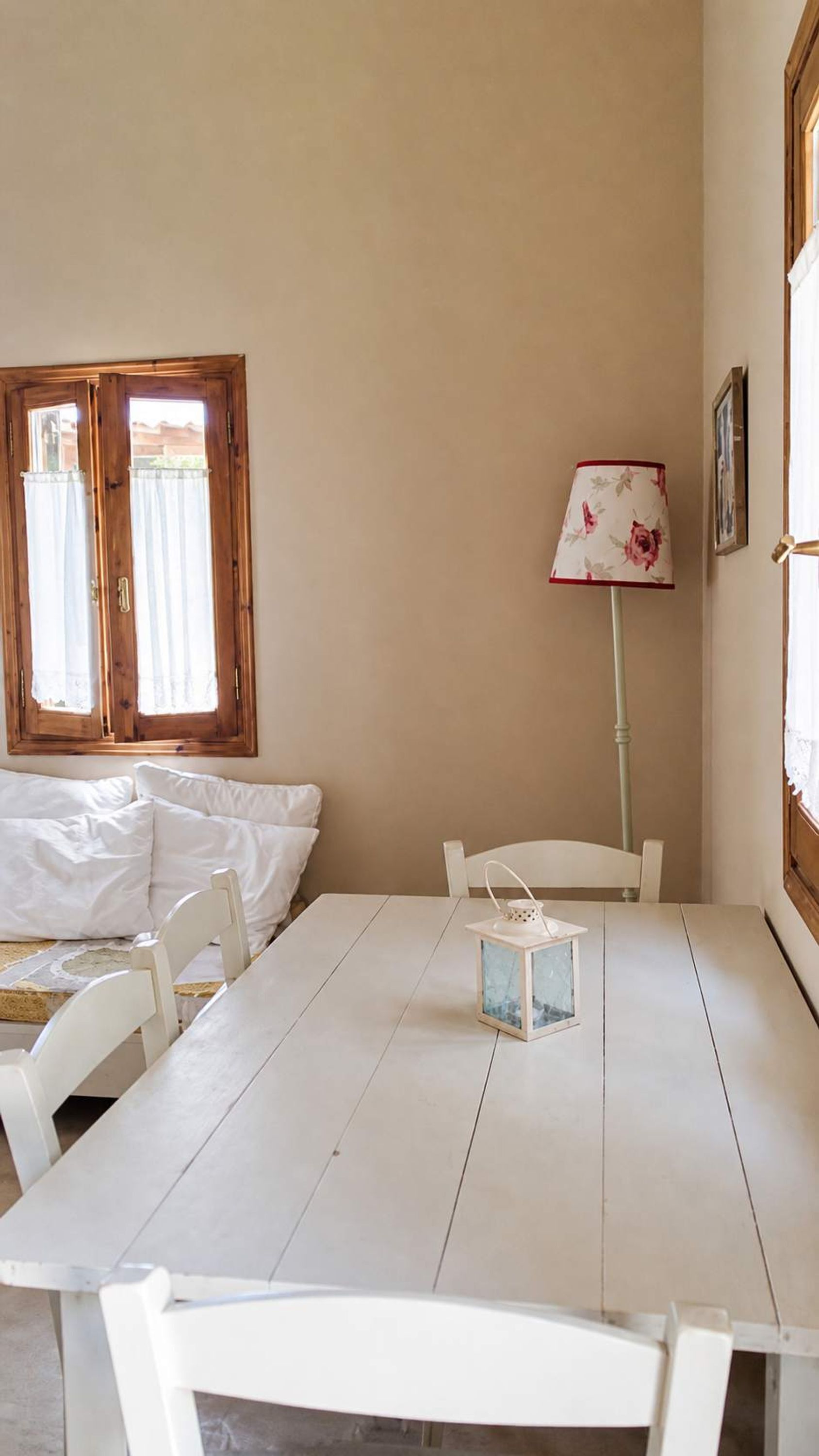 Una habitación con una mesa blanca, sillas, una cama, una ventana con cortinas y una lámpara.