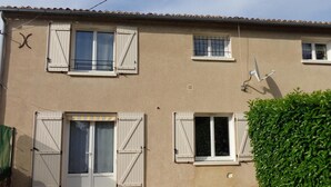 Un bâtiment à deux étages doté d’une terrasse en bois, de volets aux fenêtres et d’une haie verte.