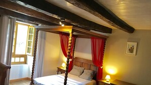 Een hemelbed met een houten hoofdeinde, twee nachtkastjes met lampen, een raam met gordijnen en een ingelijste foto aan de muur.