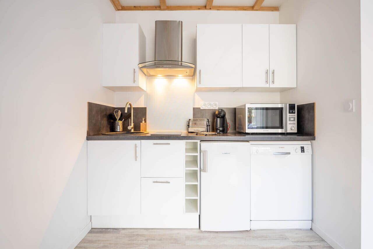 A compact kitchen with white cabinets, a stainless steel range hood, a microwave, a coffee maker, and a dishwasher. - Le Gite de l'Olivier (MONTAMISE)