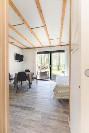 A room with a wooden ceiling, a bed, a TV, a table with chairs, and a sliding glass door leading to an outdoor area. - Le Gite de l'Olivier (MONTAMISE)