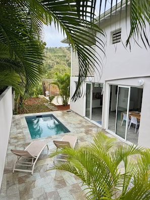Área da piscina com espreguiçadeiras, um espaço de refeições com cadeiras brancas e vista para uma encosta.