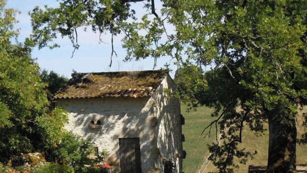 Un bâtiment rustique en pierre, doté d’un toit de chaume, entouré de verdure et d’arbres.