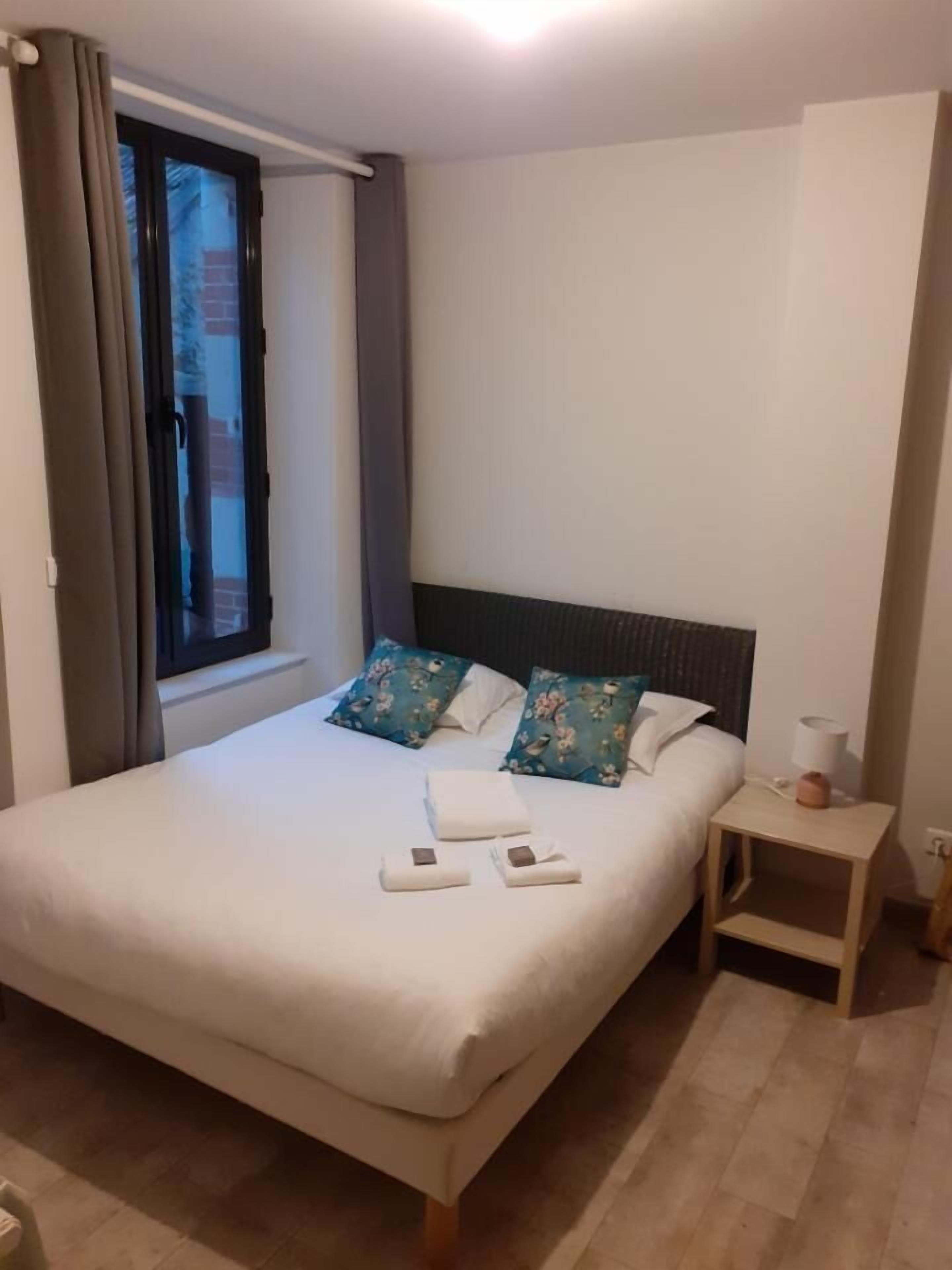 Een opgemaakt bed met bloemenkussens, een nachtkastje met een lamp en een raam met gordijnen.