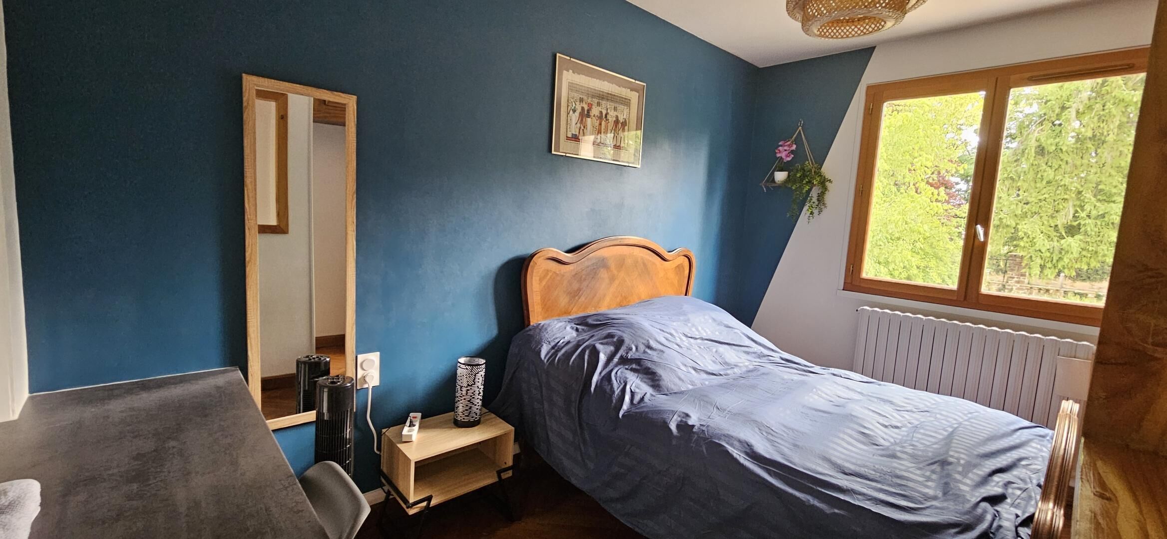 Une chambre à coucher comprenant un lit, une table de chevet, un miroir, une fenêtre donnant sur un espace verdoyant et un radiateur.